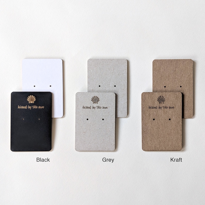 May include: Three earring display cards with a seashell and the text "kissed by the sun". The cards are black, grey, and kraft paper.