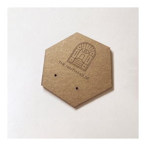 May include: A hexagonal brown cardboard tag with two holes. The tag features a line drawing of an arched doorway with an eye inside and the text "THE NINTH HOUSE".