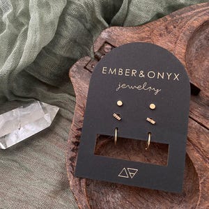 May include: A set of gold earrings on a black card with the text "Ember & Onyx Jewelry". The set includes a pair of small hoops, a pair of small studs, and a pair of small bar earrings.