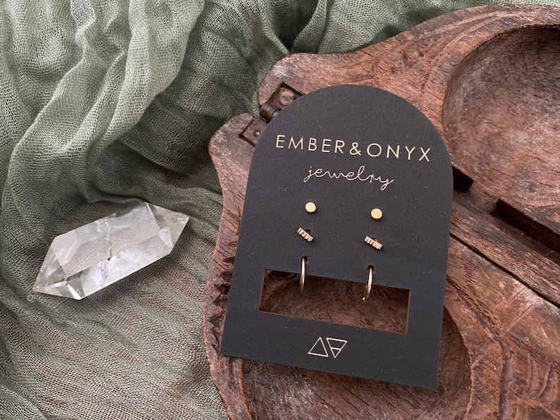 May include: A set of gold earrings on a black card with the text "Ember & Onyx Jewelry". The set includes a pair of small hoops, a pair of small studs, and a pair of small bar earrings.