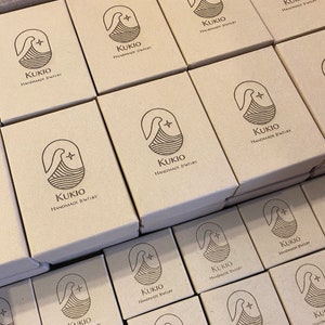 Peut inclure: Une pile de bo&icirc;tes en carton brun avec le logo "KUKIO HANDMADE JEWELRY" imprim&eacute; sur chaque bo&icirc;te.