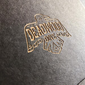 May include: A brown paper card with a stylized image of a raven with the text "Deadwood Dove" in a vintage font.