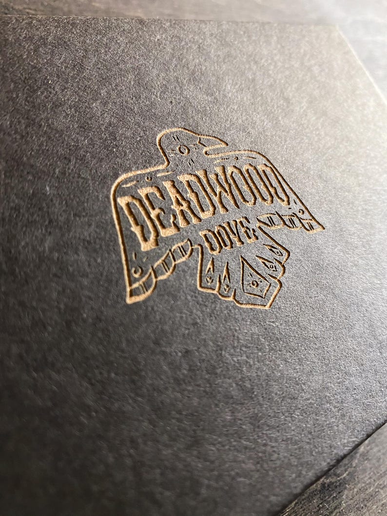 May include: A brown paper card with a stylized image of a raven with the text "Deadwood Dove" in a vintage font.