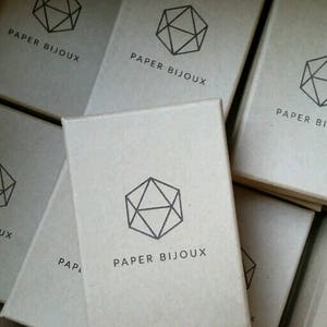 Peut inclure: Une pile de petites bo&icirc;tes en carton brun avec un motif g&eacute;om&eacute;trique et le texte "PAPER BIJOUX" imprim&eacute; sur chaque bo&icirc;te.