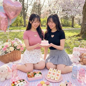 May include: Two young women are seated on a picnic blanket, holding a small pink cake. A basket of flowers, balloons, and stuffed animals surround them. The picnic includes sandwiches, snacks, and drinks. The scene is set outdoors.