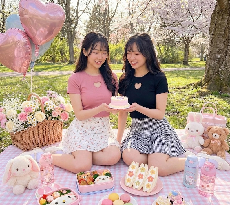May include: Two young women are seated on a picnic blanket, holding a small pink cake. A basket of flowers, balloons, and stuffed animals surround them. The picnic includes sandwiches, snacks, and drinks. The scene is set outdoors.