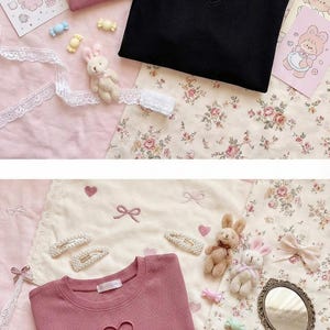 May include: Two ribbed knit shirts, one dusty rose and one black, each with a heart-shaped cutout. The shirts are displayed with decorative items like hair clips, bows, and small stuffed bunnies, creating a cute, feminine aesthetic.