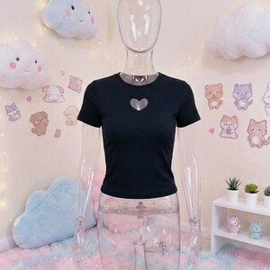 May include: Black short-sleeved crop top with a heart-shaped cutout. The top is displayed on a clear mannequin. The background features cloud-shaped pillows, stickers of cartoon animals, and a small white shelf with potted plants.