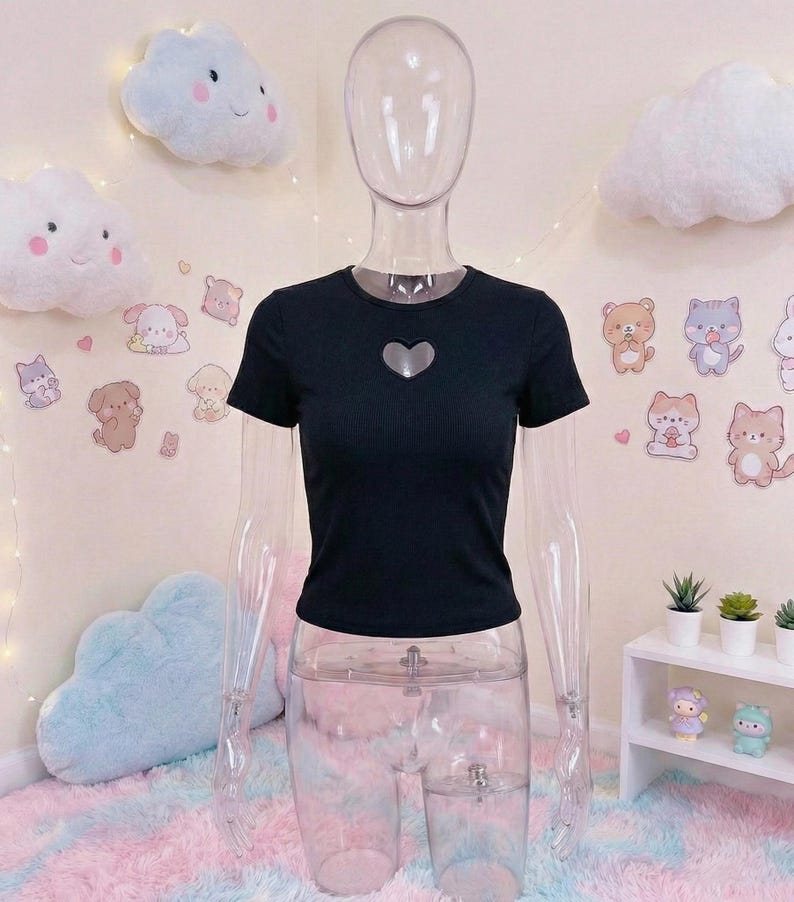 May include: Black short-sleeved crop top with a heart-shaped cutout. The top is displayed on a clear mannequin. The background features cloud-shaped pillows, stickers of cartoon animals, and a small white shelf with potted plants.