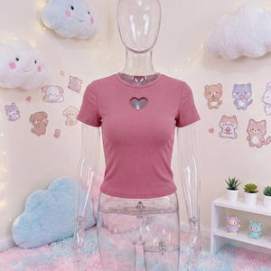 May include: A dusty rose-colored, short-sleeved top with a heart-shaped cutout at the chest. The top is ribbed and displayed on a clear mannequin. The background features cloud-shaped decorations and cartoon animal stickers.