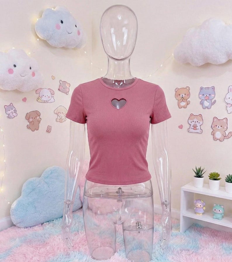 May include: A dusty rose-colored, short-sleeved top with a heart-shaped cutout at the chest. The top is ribbed and displayed on a clear mannequin. The background features cloud-shaped decorations and cartoon animal stickers.