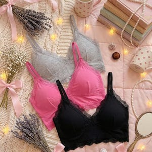 May include: Four bralettes in pink, gray, and black with lace trim are arranged on a pink surface. The bralettes are surrounded by decorative elements, including lavender, books, and mugs with heart patterns. The scene is lit with small fairy lights.