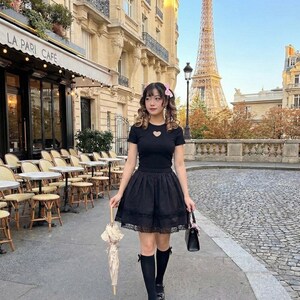 May include: A black outfit consisting of a short-sleeved top with a heart-shaped cutout and a layered skirt. The outfit is paired with black knee-high socks and shoes. The person is holding a black handbag and a cream-colored parasol. The background features a cafe and the Eiffel Tower.