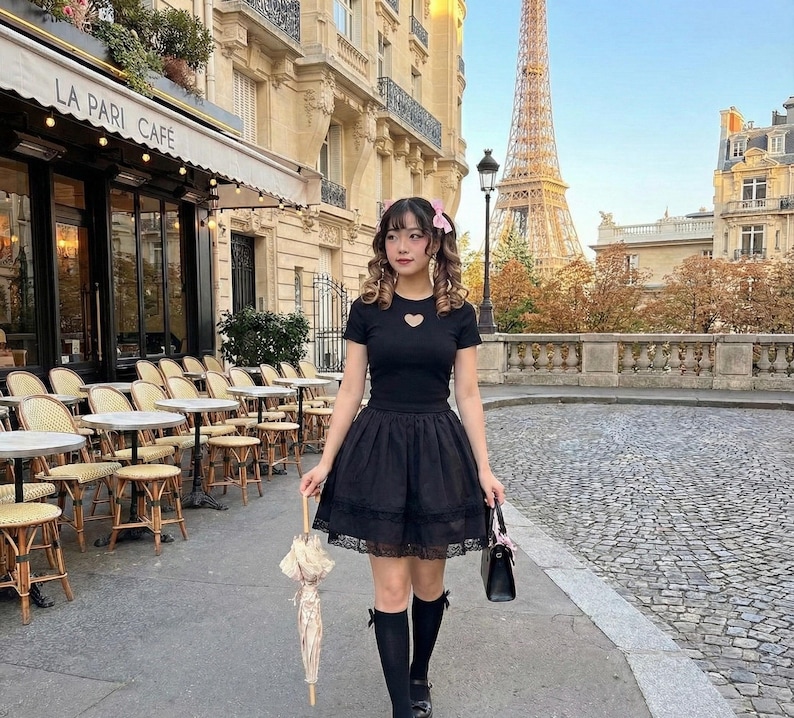 May include: A black outfit consisting of a short-sleeved top with a heart-shaped cutout and a layered skirt. The outfit is paired with black knee-high socks and shoes. The person is holding a black handbag and a cream-colored parasol. The background features a cafe and the Eiffel Tower.