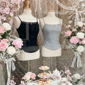 May include: A boutique window display featuring two camisole tops on mannequins, one black and one gray, both with lace trim. The display includes floral arrangements of pink and white roses, a tiered dessert stand with macarons and cakes, and a tea set.