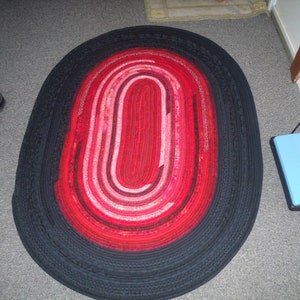 May include: A black and red oval rug with a braided design. The rug has a black outer border and a red and pink inner circle.