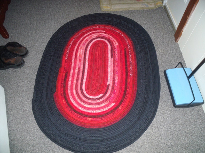 May include: A black and red oval rug with a braided design. The rug has a black outer border and a red and pink inner circle.
