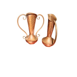 May include: Two copper vessels with a metallic finish. The left vessel features a hammered texture, a wide flared rim, and two large curved handles. The right vessel has a smooth surface, a tall cylindrical body, and a single curved handle.