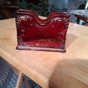 May include: A deep red glass napkin holder featuring an ornate embossed scrollwork pattern and a diamond-textured border. The center of the glass displays faint, cursive script lettering that is partially visible against the translucent surface.