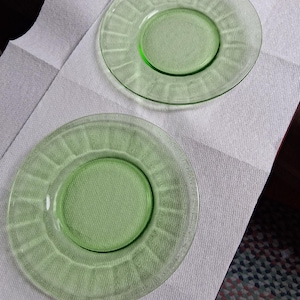 May include: Two round, translucent light green glass plates featuring a smooth center and a textured rim with a repeating vertical ribbed pattern. The plates have a clear, glossy finish and a distinct circular design radiating from the center.