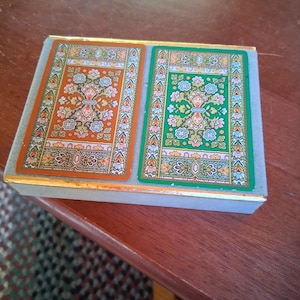 May include: A rectangular box containing two decks of playing cards. The box features a gold-toned trim and a light gray base. Each card back displays an intricate floral pattern, one with an orange background and the other with a green background.