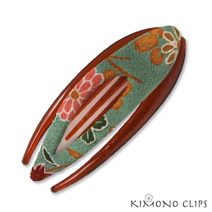 May include: A brown hair clip with a floral design. The clip has a curved shape with a green fabric center featuring orange and pink flowers. The text "KIMONO CLIPS" is at the bottom.
