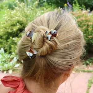 以下が含まれることがあります： ブロンドの髪の女性が、4つのべっ甲のヘアクリップを髪に付けています。クリップは頭頂部に集められ、髪をまとめたお団子になっています。