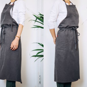 May include: A long grey linen apron with a large pocket on the front. The apron has adjustable straps and ties at the waist.