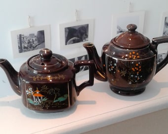 May include: Two dark brown ceramic teapots with lids, handles, and spouts. The left teapot features a painted landscape with a pagoda and bridge. The right teapot displays a floral pattern with orange, white, and blue accents on the dark glaze.