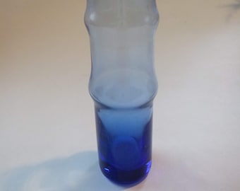 May include: A tall, cylindrical glass vase featuring a distinct bamboo-like segmented design. The glass transitions from a deep, saturated cobalt blue at the rounded base to a lighter, translucent sky blue hue toward the open, flared top rim.