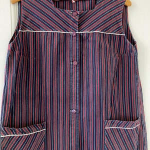 May include: A sleeveless, button-up dress with a striped pattern in red, green, and black. The dress has two pockets on the front and a white trim around the neckline and pockets.