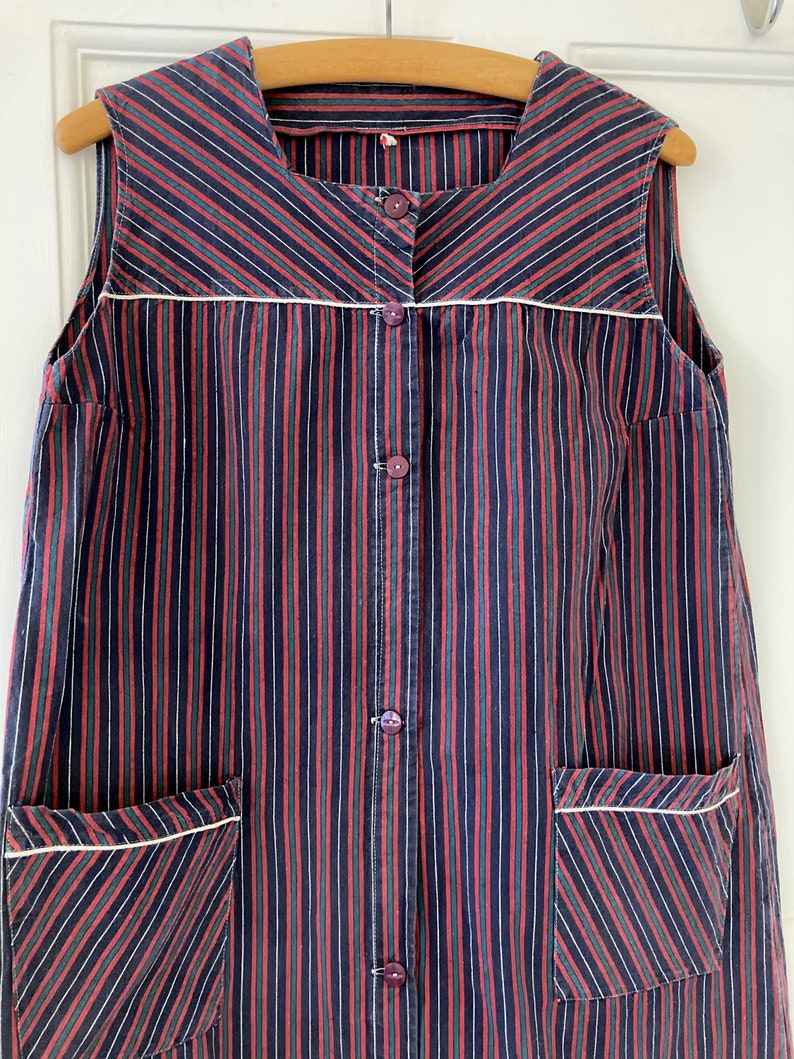 May include: A sleeveless, button-up dress with a striped pattern in red, green, and black. The dress has two pockets on the front and a white trim around the neckline and pockets.
