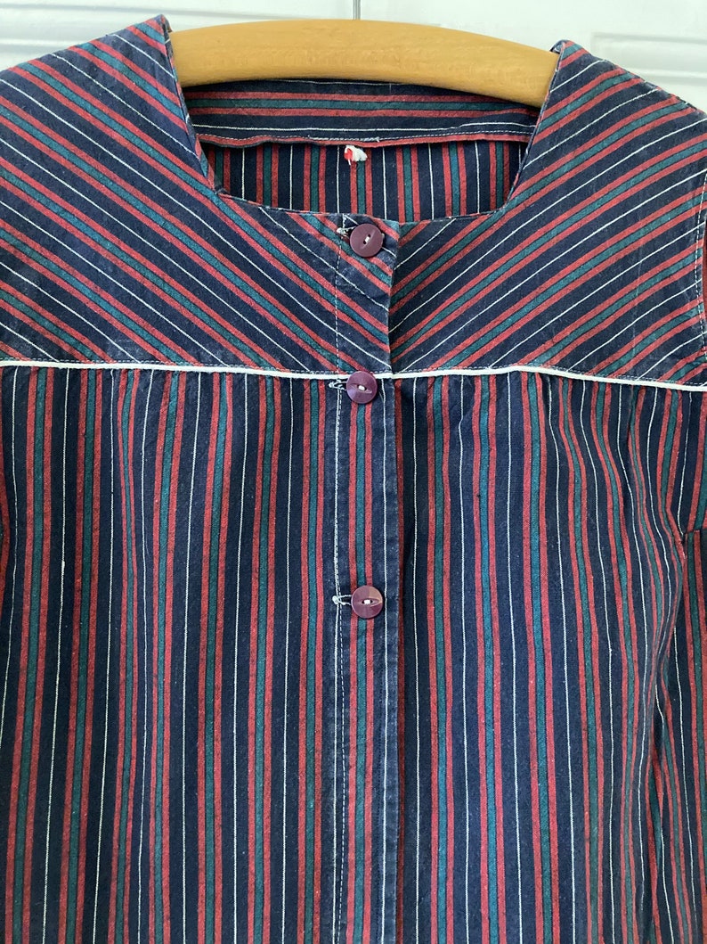 May include: A striped shirt with a white trim around the neckline. The shirt has red, green, and black stripes. The shirt has a button closure.