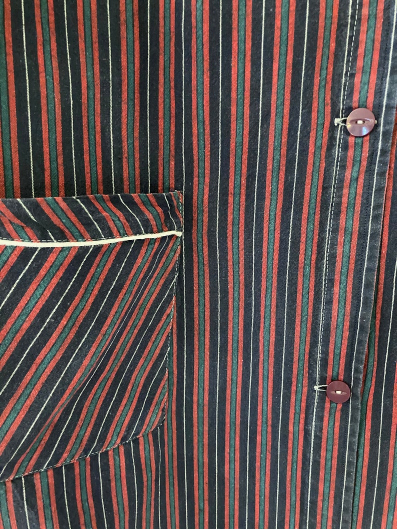 May include: A close-up of a black, red, and green striped fabric with a white pocket. The fabric has a subtle diagonal pattern. Two dark brown buttons are visible on the right side of the image.