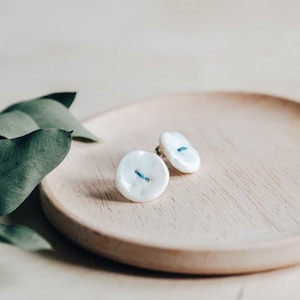 Puede incluir: Un par de pendientes de botón de cerámica blanca con un detalle de línea azul. Los pendientes están sobre una superficie de madera.