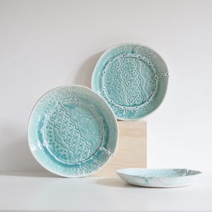 Può includere: Tre piatti in ceramica di varie dimensioni. I piatti sono di colore turchese chiaro con un design strutturato e in rilievo. I piatti sono disposti su una superficie bianca con un blocco di legno sullo sfondo.