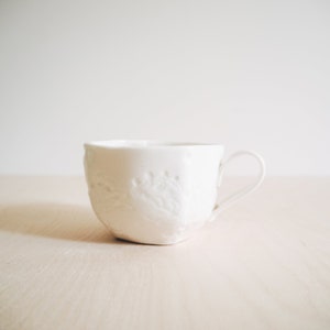 May include: A white ceramic teacup with a raised floral design. The cup has a rounded shape and a handle.