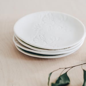 May include: Four white ceramic plates with a floral pattern, stacked on top of each other. The top plate displays the embossed floral design. The plates are on a light-coloured wooden surface. The plates are likely for serving food.