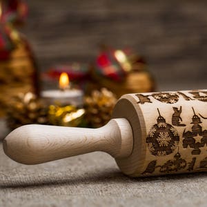 May include: A wooden rolling pin with a Christmas design. The rolling pin is engraved with images of ornaments, cats, and stars. The rolling pin is on a burlap surface.