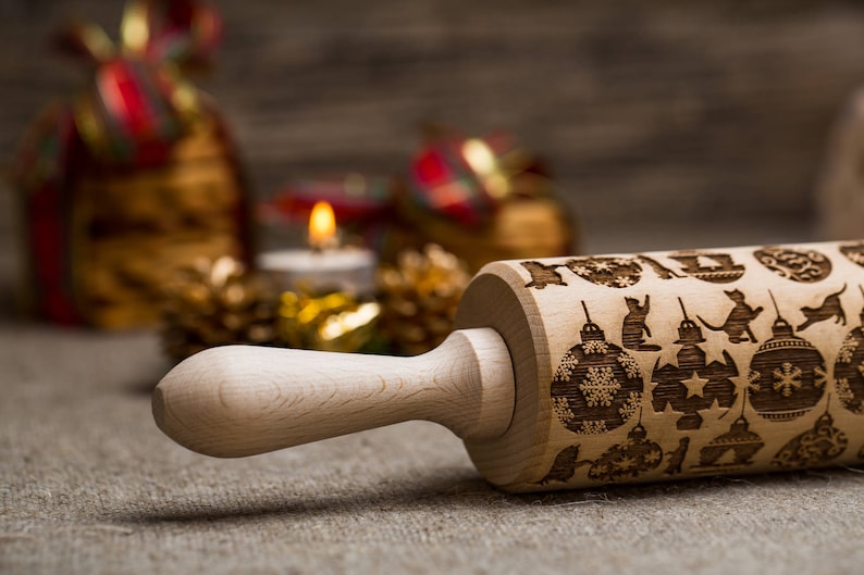 May include: A wooden rolling pin with a Christmas design. The rolling pin is engraved with images of ornaments, cats, and stars. The rolling pin is on a burlap surface.