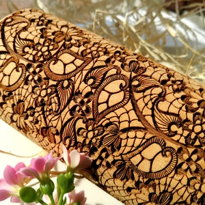 Può includere: Un mattarello di legno con un motivo a pizzo dettagliato e intricato inciso sulla superficie. Il motivo è marrone scuro contro il legno marrone chiaro.