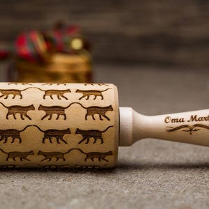 May include: A wooden rolling pin with a pattern of cats engraved on the surface. The handle is engraved with the text "Oma Marta".