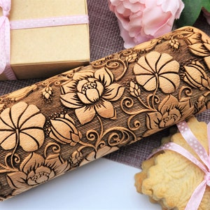 May include: A wooden rolling pin with a floral design engraved on the surface. The design features intricate details of flowers and leaves. The rolling pin has a wooden handle with the word "Oma" engraved on it.