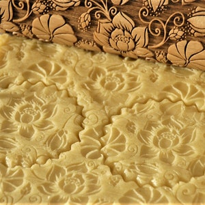 Pode incluir: Um close-up de um rolo de massa de madeira com um design floral sendo usado para abrir a massa. A massa está impressa com o design floral do rolo.