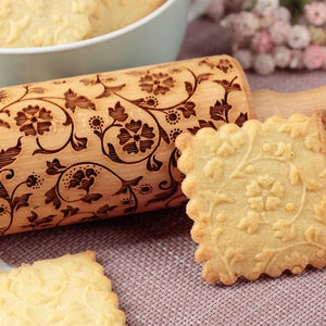 Peut inclure: Un rouleau à pâtisserie en bois avec un motif floral gravé sur la surface. Le rouleau à pâtisserie est à côté d'un biscuit carré avec un motif floral similaire. Le rouleau à pâtisserie porte le texte " Oma Mar" gravé sur la poignée.