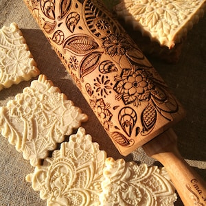 May include: A wooden rolling pin with an intricate floral design engraved on the surface. The rolling pin is used to create decorative patterns on cookies or dough. The image also shows several cookies with the same floral design.