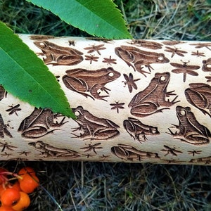 May include: A wooden rolling pin with a carved design of frogs and dragonflies. The rolling pin is light brown and the design is dark brown.