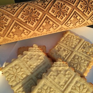 Puede incluir: Primer plano de cuatro galletas con un patrón de copo de nieve, hechas con un rodillo de madera con un diseño de copo de nieve. Las galletas son de color marrón claro y el rodillo es de color madera natural.