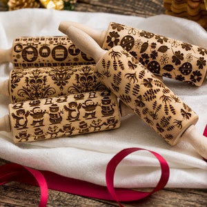 Puede incluir: Cuatro rodillos de amasar de madera con diferentes diseños grabados. Los diseños incluyen copos de nieve, un muñeco de nieve, un árbol de Navidad y un patrón floral. Los rodillos de amasar están sobre un paño blanco con una cinta roja.