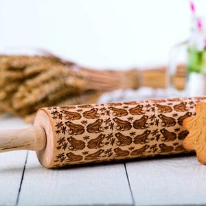 May include: A wooden rolling pin with a carved design of frogs and dragonflies. The rolling pin is on a white wooden surface with a cookie in the background.